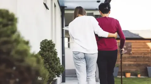 Getty Images A woman assists another, leaning on a walking stick, as they walk together past a house.