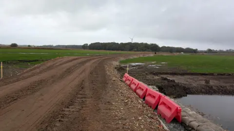 A dirt road has been created with fields either side and trees and turbines seen in the distance. Plastic red posts are at the side of the track with sandbags by water. The sky is cloudy.