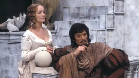 A young Frank Barrie, dressed in extravagant Shakespearean clothing, performs in a stage production of The Two Gentlemen of Verona.