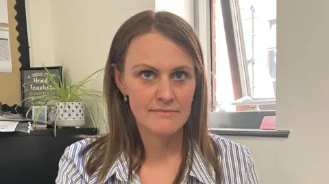 Adele Forrest/BBC A woman with long brown hair weaing a blue striped shirt sits in an office
