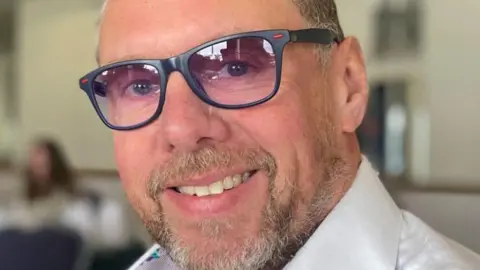 Stephen Hands, a man with short brown hair and a closely trimmed beard. He is wearing dark framed glasses that have lightly purple tinted lenses and has on a white shirt with a high collar. He is smiling.