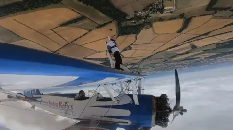 PA Media Betty Bromage wing walking. The plane is upside down in the air. The landscape of the ground can be seen, as well as the clouds in the sky.