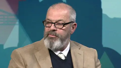 A man with short grey hair and a greying beard and wearing black-rimmed glasses and a brown suit, black pullover, white shirt and tie.