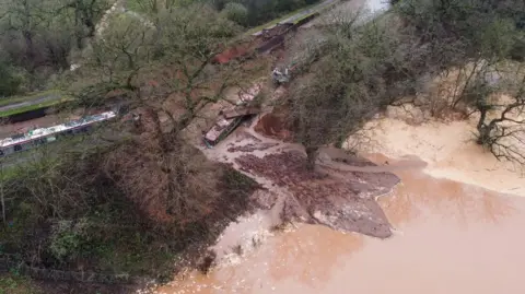 PA Media An aerial view of a large body of brown water alongside a large muddy hole with three narrow boats seen in it