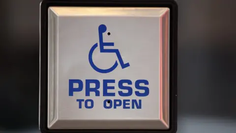 Close-up of a stainless-steel accessibility door button displaying a blue wheelchair symbol above the words “PRESS TO OPEN.” The square button is mounted on a post, with a blurred indoor background.