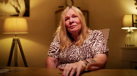 Yeti Television/BBC Cymru Wales Cathy Broomfield is a woman in late middle age. She has dyed blonde chest-length hair. She is wearing an above the elbow sleeved dress with a brown and cream patterned dress. She is seated behind a table with her left hand resting on it. She wears an large square watch and a wedding ring. She has a tattoo on her lower left arm. She is sitting in a living room with a mantlepiece to her right with a lamp on it, and a standard lamp to her right. There is an out of focus cabinet behind her and a painting on the wall.