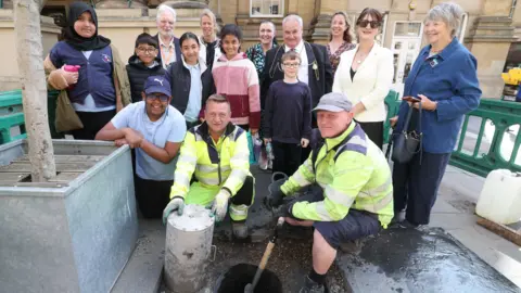 Two men wearing hi-vis yellow jackets hold a spade and a large metal cylinder as they crouch down next to a hole in concrete. A group of children and adults stand behind them looking at the camera. 