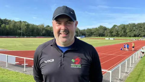 Close up picture of Barry smiling looking at the camera. Behind him is an athletics running track with a group of non identifiable children in the background. Barry is wearing a long sleeve black sports top with Ynys Mon Island Games branding on. He is also wearing a black nike cap.. 