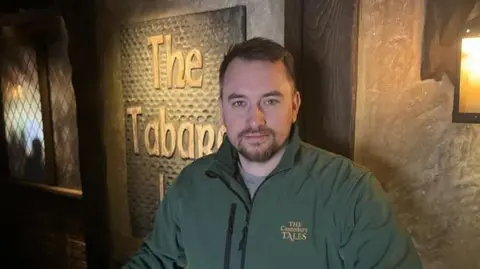 Lewis Hunt, Keeper of the Tales stands in The Tabards Inn section under low lighting in The Canterbury Tales