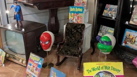 A selection of old children's toys and items on display. They include a Spider-Man mask, a 1960s TV, a Superman action figure and lots of books.