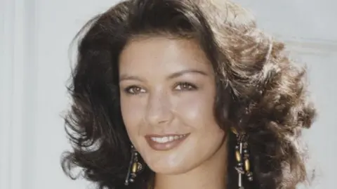 A close up of a young Catherine Zeta-Jones in character as Mariette. She has dark brown wavy hair and wears long beaded statement earrings. 