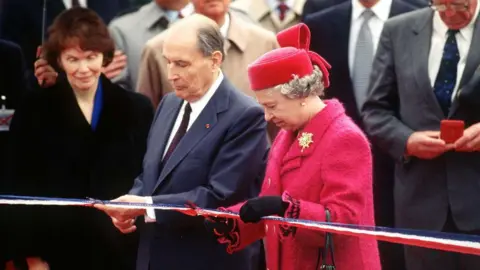 Getty/Tim Graham  President Mitterrand in a blue suit and burgundy tie stands beside Queen Elizabeth in a red dress coat and hat as they cut a ceremonial ribbon at the opening of the Channel Tunnel