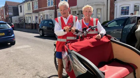 Debbie, a woman with light blond hair, and her husband, a man with grey hair, both wearing red high-visibility jackets and standing next to a bike with a passenger cart fixed onto the front, big enough to fit two people seated. They're standing in the middle of a residential street with houses and parked cars behind them.