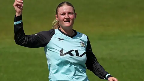 Ryana MacDonald-Gay in a light blue shirt points to the sky before bowling