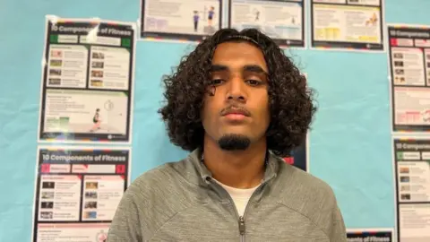 Mohammed is behind a classroom that has lots of different leaflets he is wearing a grey zipped up jacket.
