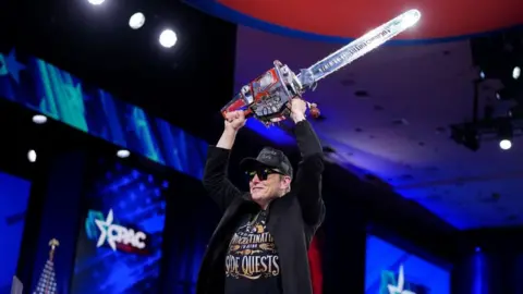 Elon Musk holds a chainsaw on stage at the Cpac conference last week, a symbol of his cutting of the federal workforce on behalf of Donald Trump.