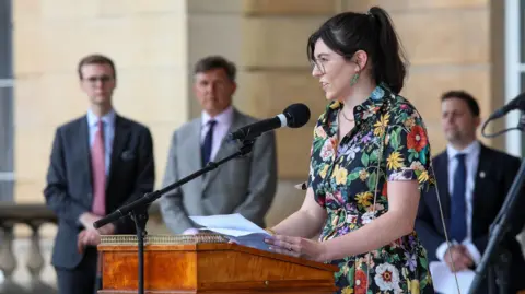 Charlotte Kemp with dark hair and glasses, wearing a multicoloured floral dress and holding sheets of paper, which are resting on a lectern. She is speaking into a microphone on a stand watched by three men in suits and ties. The light-coloured stone of Buckingham Palace is visible behind them.