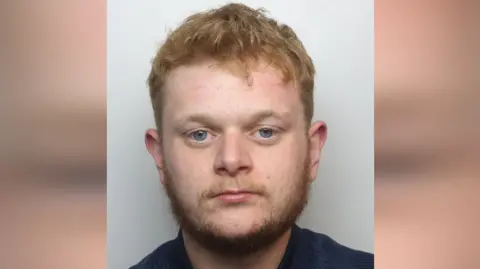 Thames Valley Police Jordan Clarkson's mugshot. He has short, ginger hair and a wispy beard. He is wearing a blue top in front of a grey background.