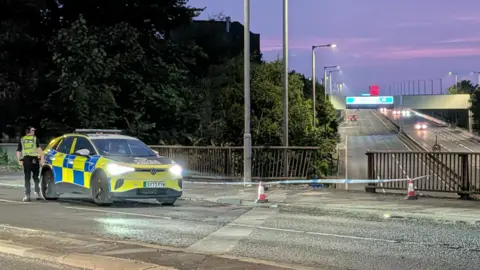 A police car is parked by police tape which is across a damaged barrier over a motorway in the evening