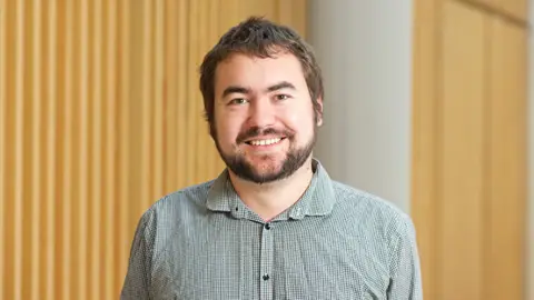 NOTTINGHAM TRENT UNIVERSITY Dr Theo Hughes‑Riley has brown hair and beard, standing in a grey shirt