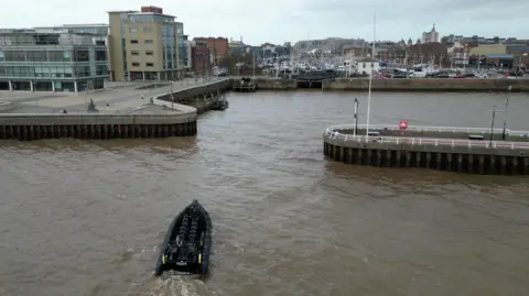 A black police rigid‑hulled inflatable boat moves towards a gap between two quayside walls. Several officers in dark protective gear sit and stand on board. On the far bank, modern glass‑fronted buildings sit beside older brick structures, with a large marina filled with masts visible further in the background. The sky is grey and overcast, and the riverfront walkways appear quiet.