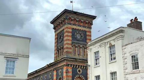 A tower without any clocks in the walls. Pigeons are flying around it. The tower is topped with a weather vane. Two smaller buildings, rendered cream, can be seen either side of the tower.