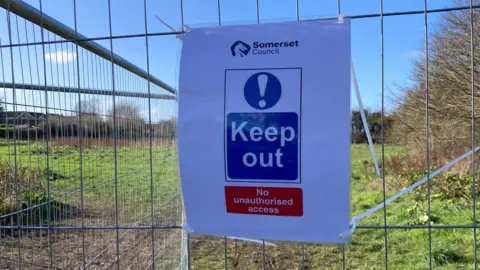 A laminated A4 sign reading: Somerset Council, keep out, no unauthorised access. It is cable-tied to a metal fence in front of a field.