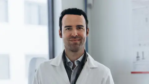 Verley Staffen Mac Milan está vestindo um casaco de laboratório branco e rindo da câmera.