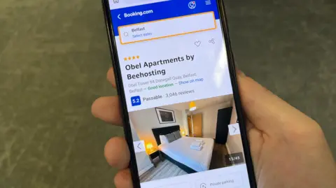 An image of a hand holding a mobile phone screen which shows the Obel apartments being advertisted on the Booking.com travel website