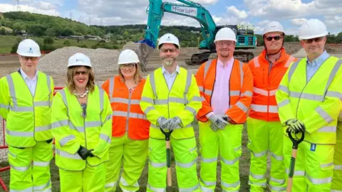 A group of five men - including Jonathan Reynolds and Jon Pearce - and two women stand smiling in high-vis yellow workwear. They all wear hard hats and hold shovels. An excavator is behind them. 