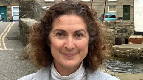 BBC Woman with shoulder length brown hair standing in front of stone bridge over river 