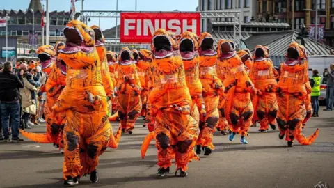 picsbycorraste A group of people in orange T. rex costumes running a long the prom with a banner that says finish in the background