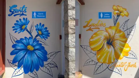Close-up of two painted walls by public toilets side by side. The left door is labelled “Gents Hommes” and decorated with large blue flowers; the right door is labelled “Ladies Dames” and decorated with large yellow flowers. The artwork is painted directly onto the wall.
