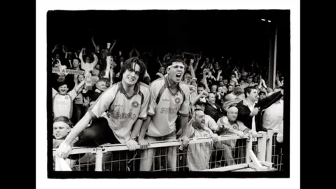 Marcus Davies Black and white photograph of two Torquay United fans looking like they are about to jump over the barriers as the fans celebrate being promoted to the old Second Division in 2004.