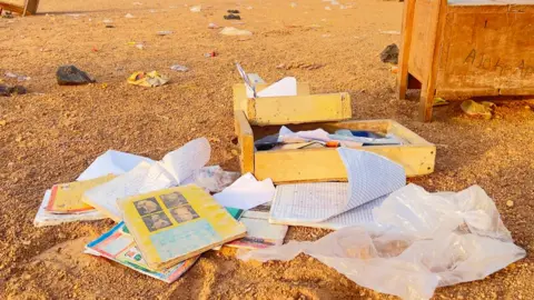 School exercise books and loose papers strewn on sandy ground flutter in the wind.