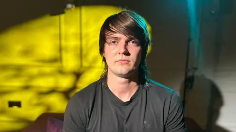 A man with shoulder-length dark brown hair looks at the camera. He is wearing a dark top