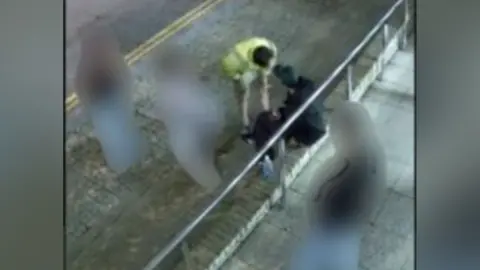 A CCTV image, taken from above, of the back of a man sitting on a low brick wall in front of some metal railings. Another man is standing and leaning forward so that his head is slightly above the other man, but close to his face. He is wearing a yellow high-vis jacket. Three other people in the image have been blurred out - two are standing next to the the high-vis man on a pavement next to a road; the other is standing on a paved area on the other side of the metal railing.