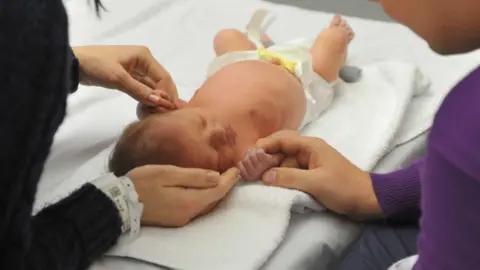 A newborn baby in a nappy has his hands held by both of his parents as he lies on a clean white towel in hospital.