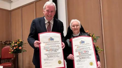 BBC/Ed Hanson Paul Callaghan stands next to Sister Mary Scholastica as they hold up their ceremonial scrolls which they recieved when they got Freedom of the City of Sunderland. 