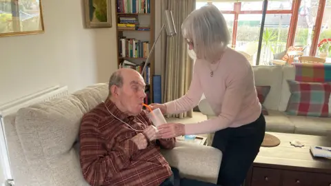 Alex Pope/BBC Clive is being given some water by his wife Barbara. They are in a living room, with two sofas. Clive is sitting down, wearing a brown-checked shirt, Barbara is wearing a pink top and black trousers. A book case stands to the left and a window is behind them. 