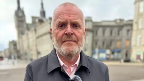 Ron Constable looking straight at the camera. He has short fair hair and a short fair and white beard. He is wearing a black jacket over a pink shirt and is standing in front of a blurred outdoor background with liht-coloured buildings.