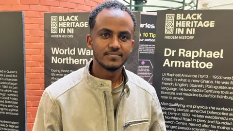 Mutwakil Bella is in a cream jacket. He has short black hair. 
He is standing in front of two museum placards. 