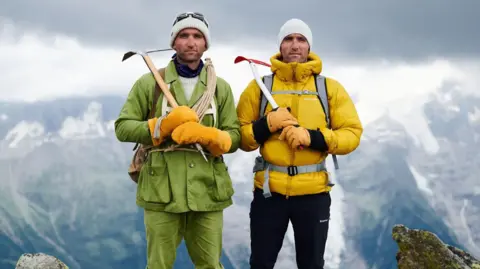 The Turner Twins Hugh and Ross standing with a backdrop of mountains, wearing their kit. Hugh's kit is a swampy green colour, and he is wearing two large yellow gloves. Ross' kit is much more modern. He is wearing a yellow puffer jacket and navy trousers.