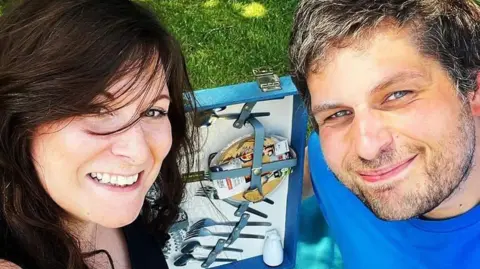 Paul Wassell Paul Wassell is right on the photo, he is looking up at the camera and is wearing a blue T-shirt. His partner Karen is on the left and also looking up at the camera and has dark hair falling into her face. They are sat next to an open picnic hamper.
