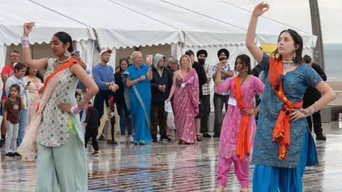 Caroline James Photography Three young female Indian dancers dancing on the comedy carpet in Blackpool, with their right arms raised and people watching in the background in front of three white marquees.