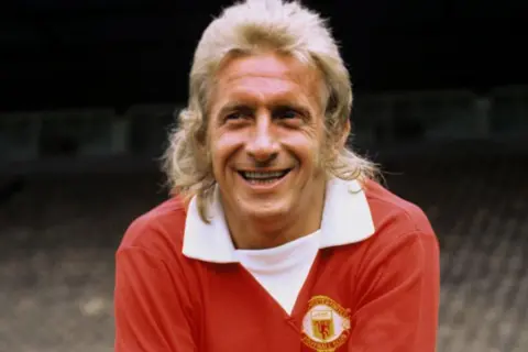 PA Media Denis Law, with a blonde mullet, in his early 1970s Manchester United red shirt with extended white V-necked collar