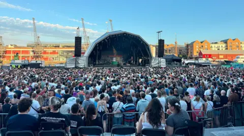 A wide shot of the stage and crowd at Bristol Sounds Festival during the evening. The sun is setting on the buildings opposite the river and the sky is blue with wispy clouds.