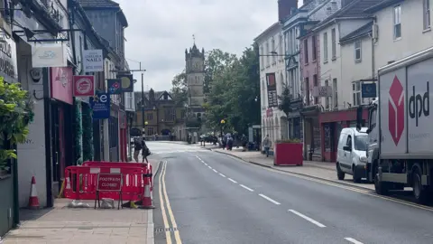 Chris Taylor Red barriers blocking off a pavement by yellow lines and a clean, new-looking road with shops around it.