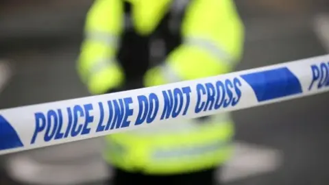 Police officer stands behind police tape reading police in blue on white background