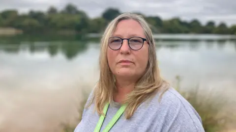BBC Councillor Brady Hughes is pictured in front of Ensor's Pool. She has shoulder length light blonde hair and is wearing back round glasses, a grey shirt and green lanyard.